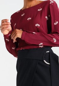 Blouse bordeaux à imprimé floral blanc, manches longues et encolure asymétrique ; associée à un pantalon foncé avec ceinture à anneau argenté.