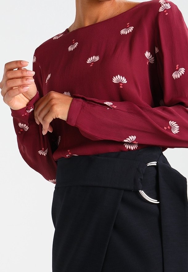 Blouse bordeaux à imprimé floral blanc, manches longues et encolure asymétrique ; associée à un pantalon foncé avec ceinture à anneau argenté.