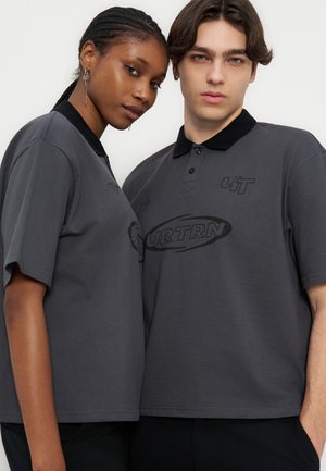 Δύο νέοι που φοράνε ίδια σκούρα γκρι πόλο μπλουζάκια με μαύρους γιακάδες, στέκονται κοντά ο ένας στον άλλο και κοιτούν μπροστά, με φόντο ένα απλό σκηνικό.