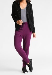 Sudadera negra con cremallera sobre una camiseta sin mangas gris, combinada con pantalones jogger morados y zapatillas deportivas negras, mostrando un estilo atlético casual.