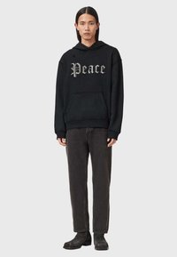 Homme aux cheveux longs portant un sweat à capuche noir avec le texte "Peace", un jean gris foncé et des bottes noires, debout devant un fond clair uni.