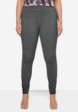 Kvinde iført tætsiddende mørkegrå leggings med blondeudskæringsdetaljer ved anklerne, kombineret med en farverig mønstret top og åbne hæle.