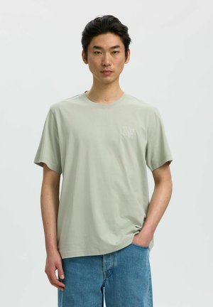 Junger Mann mit kurzen schwarzen Haaren, der ein hellgraues T-Shirt mit kurzen Ärmeln und blaue Jeans trägt, steht vor einem schlichten weißen Hintergrund.