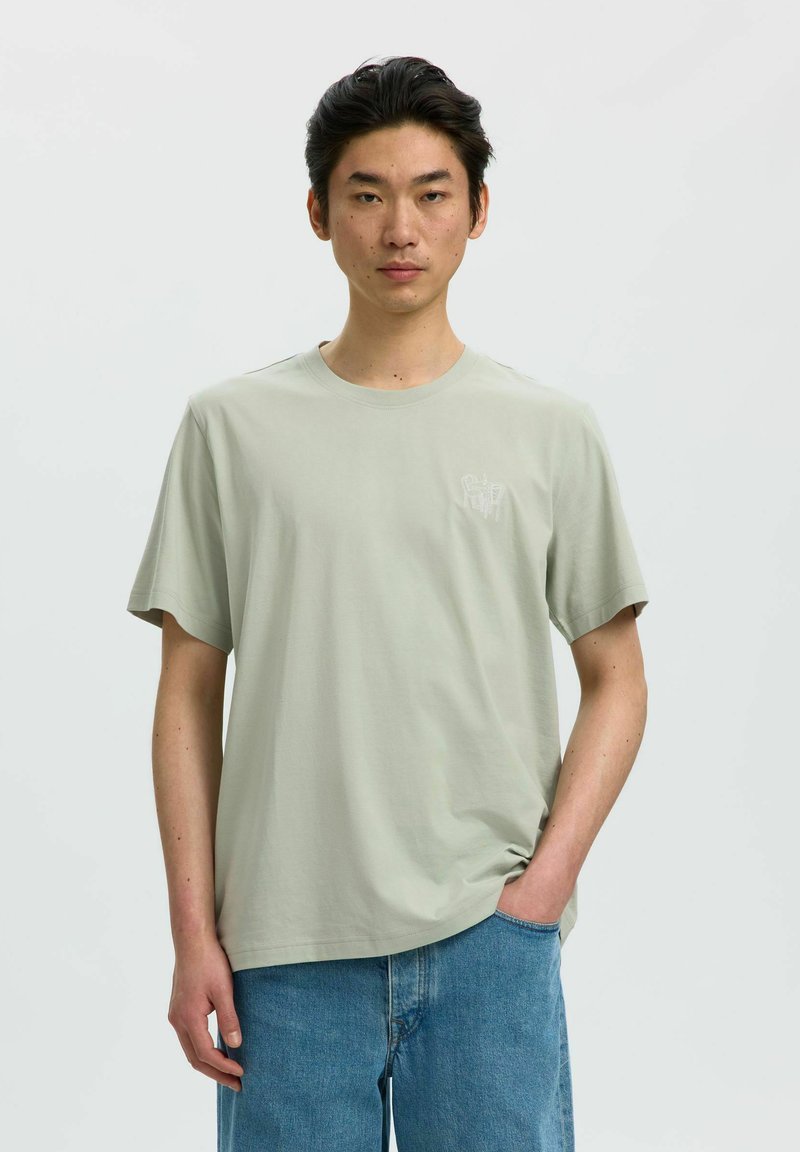 Jeune homme aux cheveux noirs courts portant un t-shirt gris clair à manches courtes et un jean bleu, debout devant un fond blanc uni.