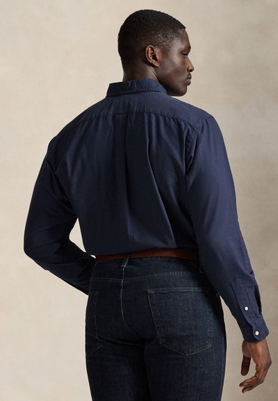 Vyras, stovintis nugara, dėvintis tamsiai mėlyną ilgomis rankovėmis marškinius įkištus į tamsiai mėlynus džinsus su ruda diržu ant vientisos spalvos fono.