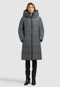 Grå pufferjakke med stort hætte, horisontal quiltet design, frontlynlås og sidelommer. Stylized over sorte tights og stiletter.