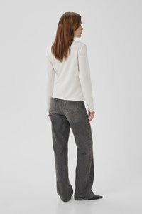 Femme aux cheveux raides roux foncé, vue de dos, portant un haut blanc à manches longues, un jean gris taille haute et des chaussures noires à bouts pointus.