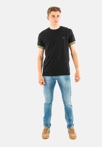 Zwarte t-shirt met korte mouwen en gele gestreepte boorden, vergezeld van lichtblauwe jeans en tan sneakers.