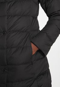 Musta toppatakki, jossa tikattu design, napillinen suljenta ja sivutasku. Sileä pinta, polven pituinen ja vartalonmyötäinen siluetti.