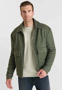 Chaqueta verde texturizada con cuello y dos bolsillos frontales, combinada con una camisa clara. Corte ajustado, cierre con cremallera en el frente.