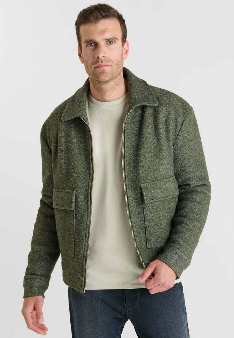 Chaqueta verde texturizada con cuello y dos bolsillos frontales, combinada con una camisa clara. Corte ajustado, cierre con cremallera en el frente.