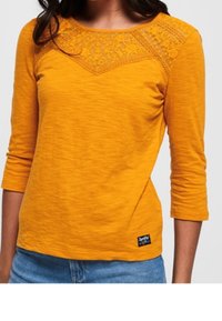 Vrouw in een mosterdgele langmouwtop met kantdetails over de bovenste borst en schouders, in combinatie met blauwe jeans.