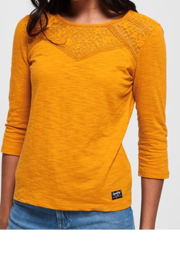 Vrouw in een mosterdgele langmouwtop met kantdetails over de bovenste borst en schouders, in combinatie met blauwe jeans.
