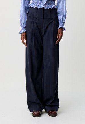 Persona che indossa pantaloni a gamba larga blu navy a righe sottili, scarpe marroni e una camicetta a righe blu e bianca con polsini arricciati.