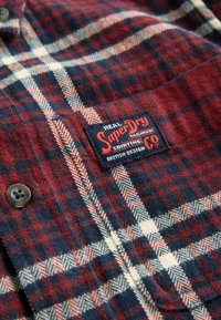 Czerwona, granatowa i kremowa tkanina w kratę z naszytą etykietą marki SuperDry oraz brązowym guzikiem wyszytym napisem SuperDry.