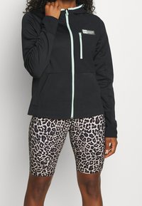 Svart zip-up hoodie med ljus turkosa detaljer, med huva och framfickor, kombinerad med leopardmönstrade cykelbyxor. Mjuk tygkänsla.
