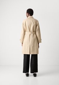 Beige trench coat s pásom, po kolená, vyrobené z hladkej látky. Má klasický golier a jemné prešívanie. Čierne široké nohavice.