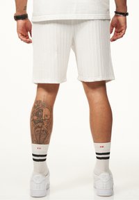 Weiße gerippte Shorts mit lockerem Schnitt, umgeschlagenen Säumen, kombiniert mit weißen Sneakers und gestreiften Socken. Tattoo am Unterschenkel sichtbar.