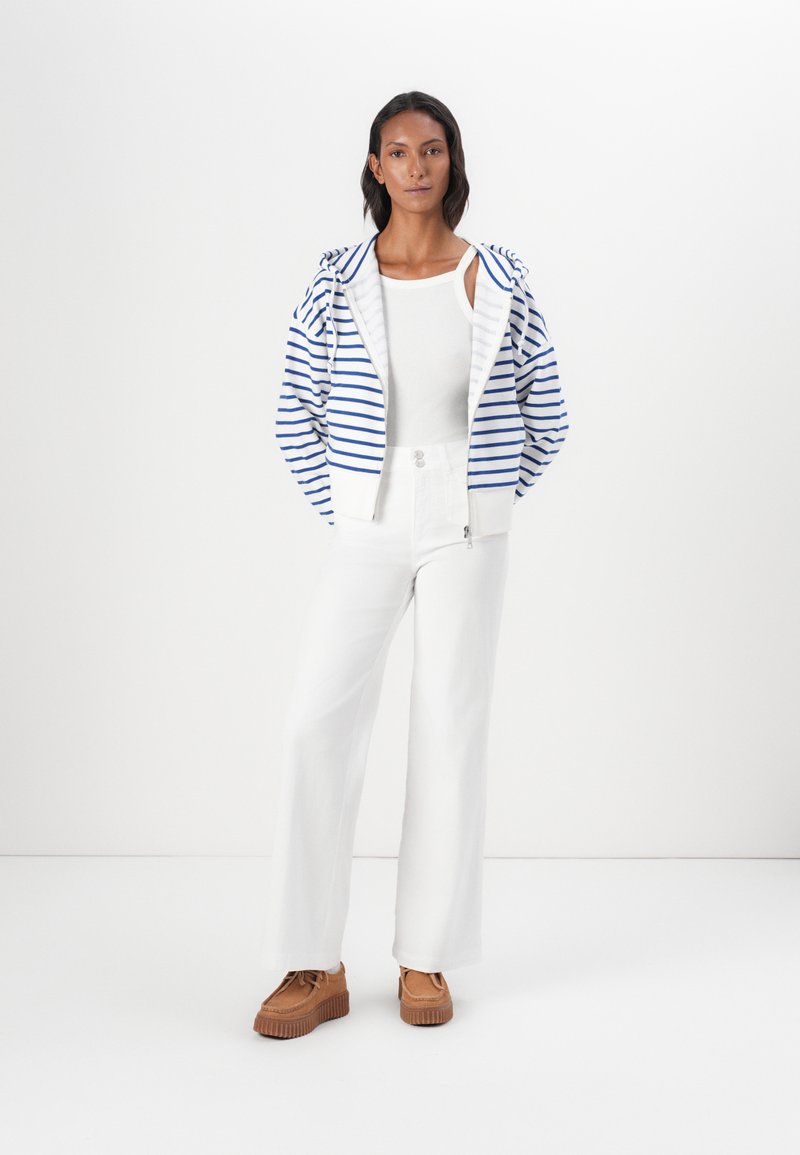 Pasiaste bluza na zamek w niebiesko-białe paski, zestawiona z białymi szerokimi spodniami i brązowymi butami na platformie. Gładkie tekstury i luźny krój.