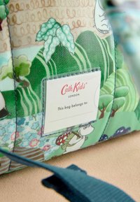 Krāsains zaļš un bežs bērnu soma ar dabu iedvesmotu rakstu, siksnu un "Cath Kids London" etiķeti ar tekstu "Šī soma pieder:".