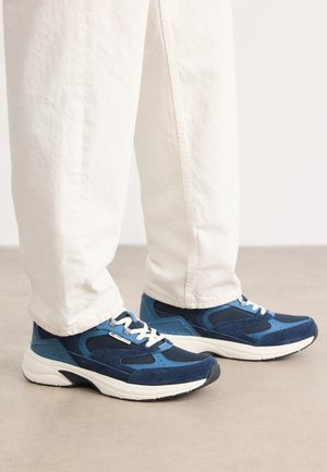 Persona vistiendo pantalones blancos y zapatillas deportivas azules con suelas y cordones blancos, de pie sobre un suelo beige.