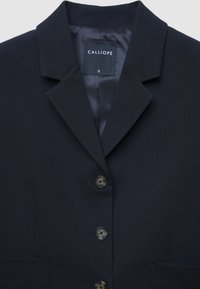 Blazer navy con colletto a punta, quattro bottoni e due tasche frontali. Tessuto liscio con fodera interna in raso. Etichetta del marchio visibile.