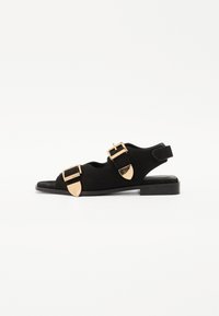 Sorte semskede sandaler med flat såle, to brede stropper pyntet med gullspenner og detaljer, åpen tå og støtte ved hælen.