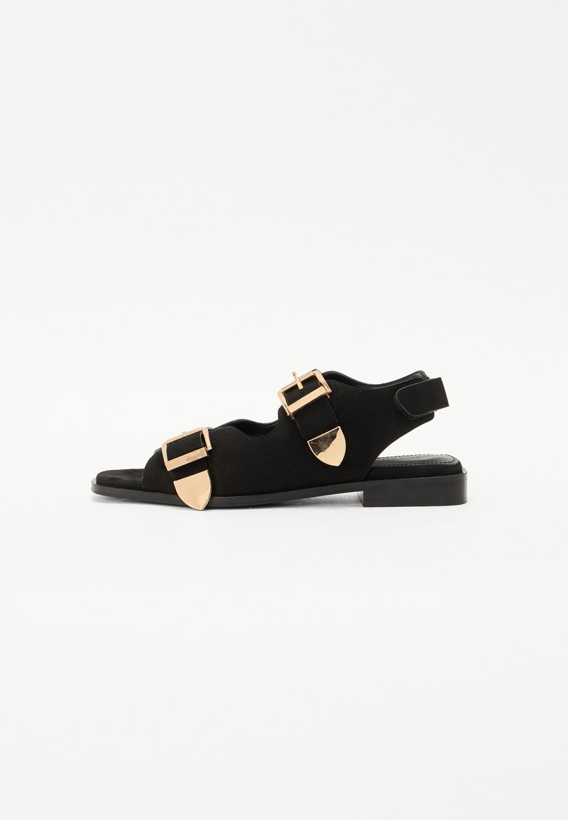Sorte semskede sandaler med flat såle, to brede stropper pyntet med gullspenner og detaljer, åpen tå og støtte ved hælen.