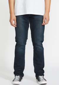 Jeans de mezclilla azul oscuro con un corte ajustado, que presentan un diseño clásico de cinco bolsillos y costuras en contraste. Combinados con zapatillas negras.
