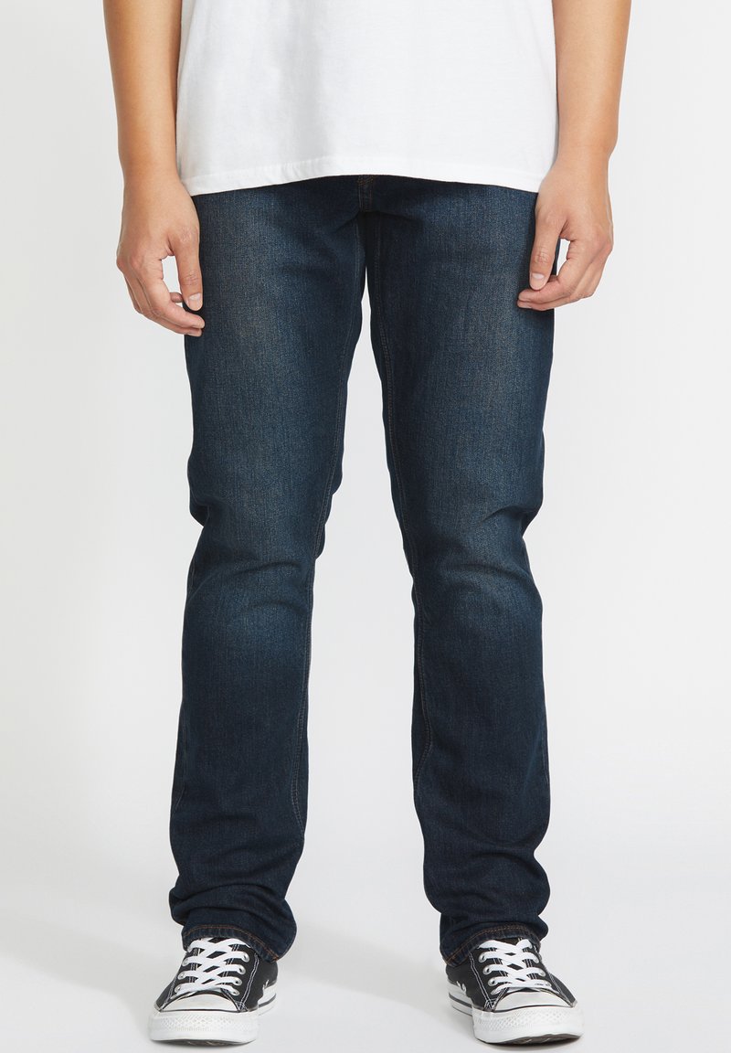 Jeans de mezclilla azul oscuro con un corte ajustado, que presentan un diseño clásico de cinco bolsillos y costuras en contraste. Combinados con zapatillas negras.