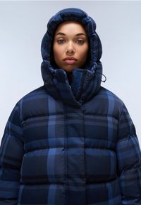 Marineblå jakke med hætte, lavet af en puf, med et ternet mønster og elastiske snore. Blød, quilted tekstur med en holdbar finish.