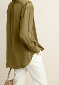 Blouse légère de couleur vert olive avec des manches longues, en tissu texturé, et des accents de dentelle décoratifs le long des épaules et des poignets. Portée avec un pantalon blanc.