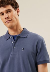 Navyblauwe polo met een gestructureerde stof, een drieknops sluiting en een klein geborduurd wit boomlogo op de linkerborst.