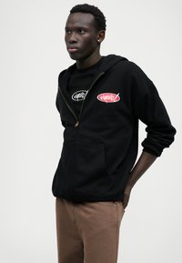 Black zip-up hoodie with embroidered logo, paired with a black t-shirt. Model wears brown pants, showcasing a relaxed fit and smooth texture.