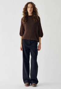 Rjav, strukturiran pleten sveter s kratkimi, širokimi rokavi. Združen z ohlapnimi črnimi kavbojkami in rjavimi čevlji, na enobarvni podlagi.