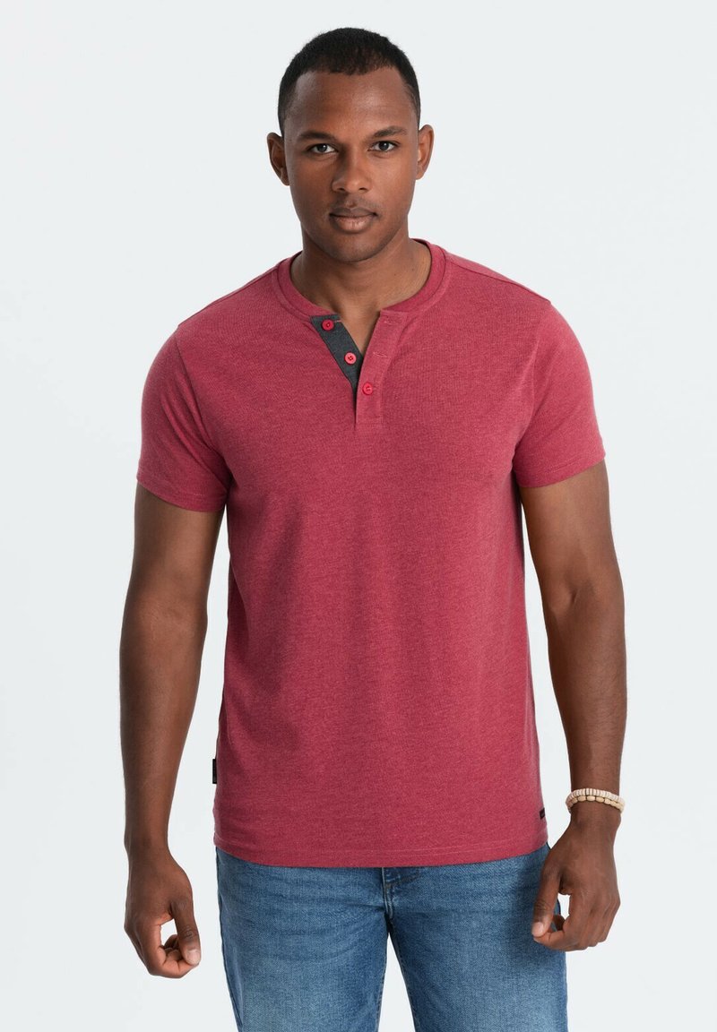 Camisa henley roja de manga corta con una tela texturizada, con una tapeta de botones en gris oscuro. Combinada con unos vaqueros azul claro.
