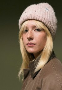 Lichtroze gebreide beanie met een textuurpatroon en een klein logo. Model draagt een bruine colbertjas, wat de zachtheid en warmte benadrukt.