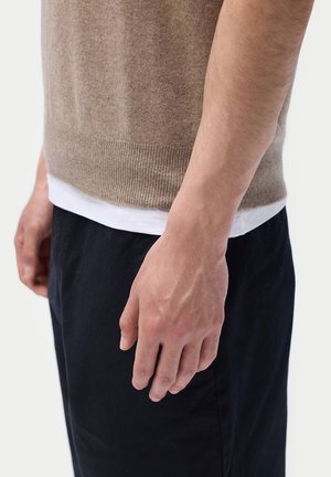 Seitenansicht einer Person, die einen braunen Pullover über einem weißen Hemd und schwarze Hosen trägt, mit einer entspannt herabhängenden rechten Hand.
