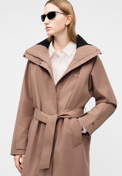 Γυναίκα που φοράει μπεζ παλτό με ζώνη και μεγάλο γιακά πάνω από λευκό πουκάμισο και μαύρα γυαλιά ηλίου, χέρι στην τσέπη, ουδέτερο φόντο.