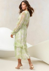Rochie florală de culoare verde deschis, din material transparent, cu mâneci lungi, tiv creț, talie strânsă, combinată cu sandale aurii cu barete, textură netedă.