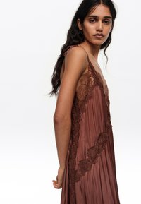 Ženska z dolgimi temnimi lasmi, oblečena v brezrokavno rjavo svileno obleko z čipkastim robom, stoji vstran na belem ozadju.