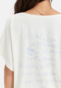 Camiseta blanca de ajuste holgado con escote ancho; presenta texto en azul claro y un gráfico de palmera en la parte posterior. Textura de tejido suave y ligero.