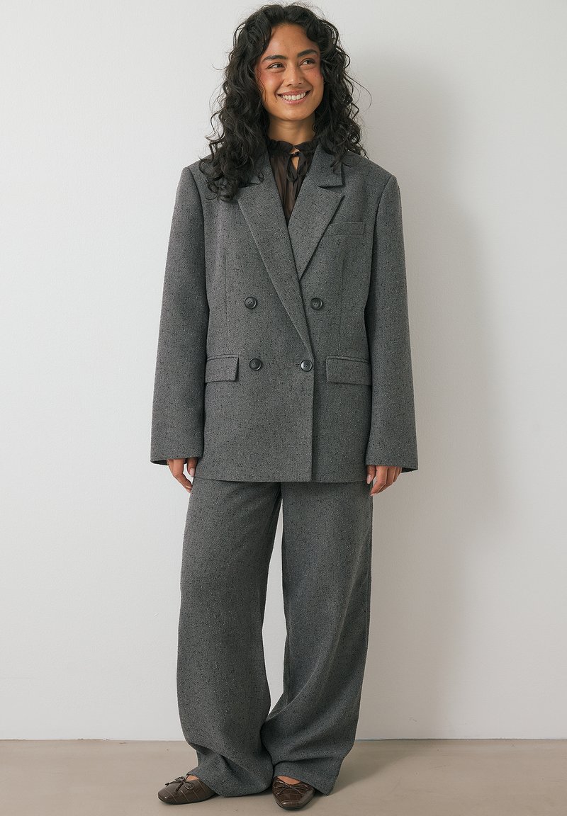 Grijze, gestoffeerde, dubbelbreasted blazer met bijpassende broek; met grote knopen, wijde pijpen en een borstzak. Klassiek op maat gemaakte stijl.
