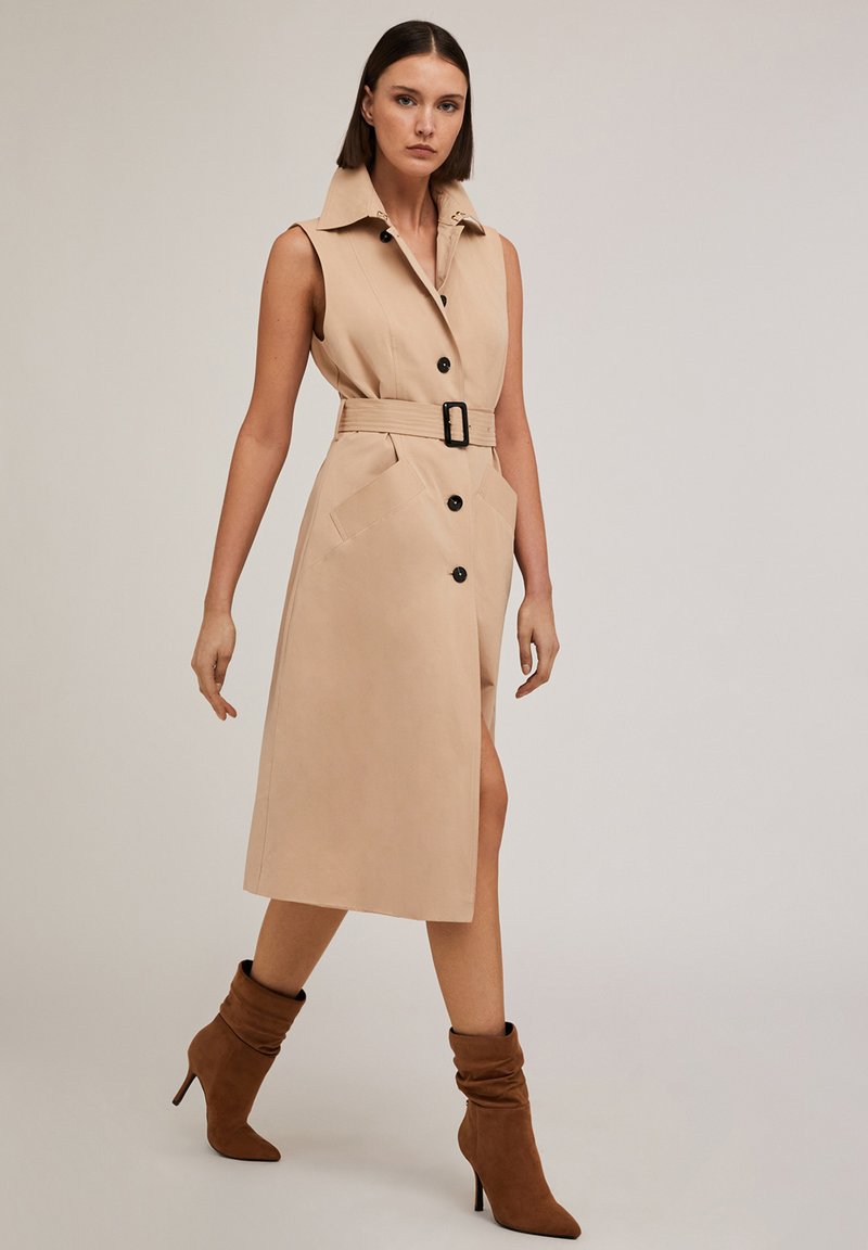 Abito trench senza maniche beige con bottoni frontali, vita con cintura e tasche laterali, abbinato a stivali con tacco marroni e morbidi.
