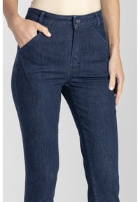 Osoba nosząca ciemnoniebieskie dżinsy z jedną ręką w kieszeni z przodu i widocznym małym tatuażem na nadgarstku.