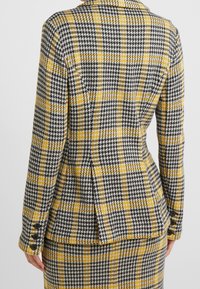 Blazer s houndstooth vzorem v černé, bílé a žluté barvě. Disponuje přiléhavým střihem, klopovým límcem, jedním zadním ventilem a knoflíkovými akcenty na rukávech.
