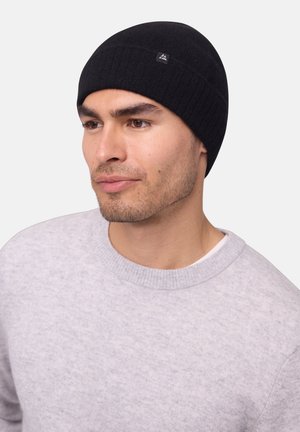 Black knitted beanie with a ribbed design and a small logo patch. Worn with a light gray crew neck sweater.