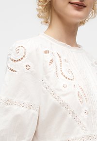 Femme aux cheveux blonds bouclés portant un chemisier blanc avec bordure en dentelle et des détails complexes de broderie ajourée sur les manches et la poitrine.