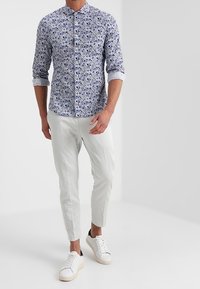 Camicia button-up blu con motivo floreale, abbinata a pantaloni bianchi a sigaretta e sneakers bianche, che mostrano un design moderno e casual.