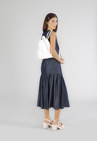 Zaino in finta pelle bianca con due zip, tasca frontale e spalline regolabili. Indossato da una modella con un vestito plissettato blu scuro e scarpe bianche.